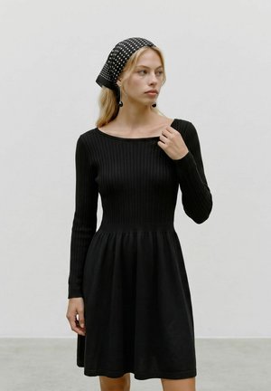 Donna con un abito nero a maglia a coste e un foulard nero a pois, in piedi davanti a uno sfondo chiaro e uniforme.