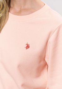 Světle růžová mikina s kulatým výstřihem, měkkou texturou a malým červeným vyšívaným logem na levé straně hrudi.