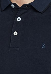 Polo bleu marine en tissu texturé, avec un col, trois boutons blancs et un petit logo brodé dans une teinte plus claire.