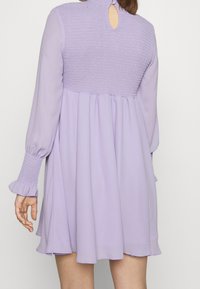 Fialové šaty se smockovaným živůtkem, dlouhými rukávy a výstřihem na zádech. Plynulá sukně s volánkovými manžetami a hladkou texturou.