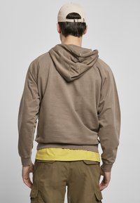 Felpa marrone con una vestibilità comoda e cappuccio con lacci. Indossata sopra una maglietta gialla, abbinata a pantaloni cargo di colore verde oliva.