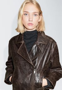 Braune Lederjacke mit großem Kragen, Frontreißverschluss und Seitentaschen. Strukturierte Oberfläche und lockere Passform betonen das Design.