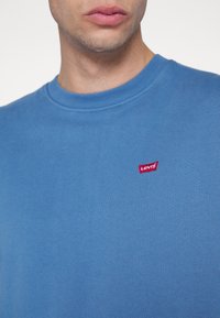 Homme portant un t-shirt Levi's bleu à col rond avec un logo Levi's rouge sur la poitrine gauche, visible du cou au haut du torse.