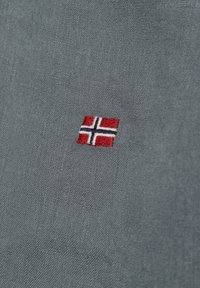 Grijze stof met een gladde textuur, voorzien van een kleine geborduurde Noorse vlag in rood, wit en blauw in de linksonderhoek.