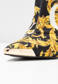Czarne materiałowe botki z złotym barokowym wzorem, spiczastym noskiem oraz metalicznym złotym akcentem na czubku z grawerunkiem "Versace Jeans Couture".