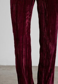 Pantalones de terciopelo burdeos triturado con una sutil textura vertical, mostrados desde el muslo medio hasta el tobillo contra un fondo neutro.