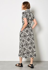Schwarzes und weißes gemustertes Maxikleid mit kurzen Ärmeln, das ein geometrisches Diamantdesign aufweist, eine Bindegürtelschlaufe hat und zwei Seitentaschen besitzt.