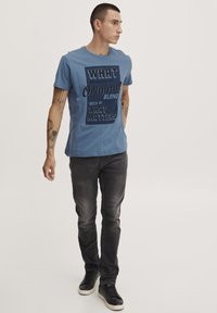 Blå bomulds T-shirt med stort sort tekst, parret med mørkegrå jeans og sorte sneakers, mod en ensfarvet hvid baggrund.