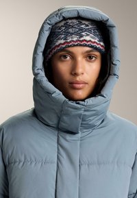 Pehmeä vaaleansininen untuvatakki, jossa korkea kaulus ja iso huppu, sileä pinta. Neulottu pipo, jossa on kuviollinen design tummansinisen ja punaisen sävyissä.