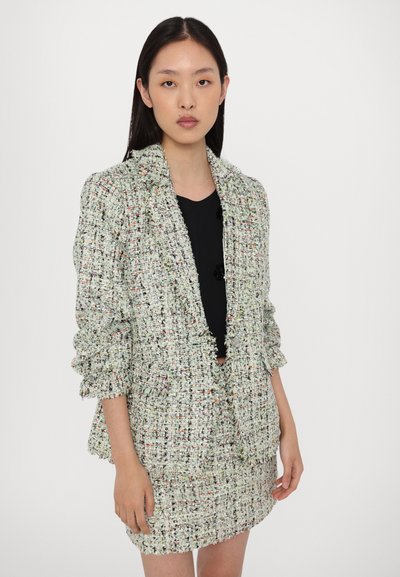 Hellfarbener Tweed-Blazer mit strukturierter Oberfläche, hochgerollten Ärmeln und lockerer Passform, kombiniert mit einem passenden Rock und einem schwarzen Oberteil darunter.