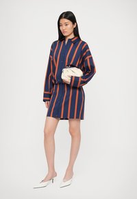 Vestido camiseiro riscado em azul marinho e laranja, com botão à frente, mangas longas, corte descontraído, combinado com uma clutch creme e sapatos de salto branco com bico fino.