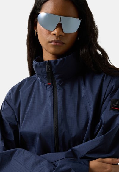 Žena s velkými zrcadlovými slunečními brýlemi, stříbrnými kruhovými náušnicemi a tmavě modrou bundou na zip, se zkříženýma rukama.