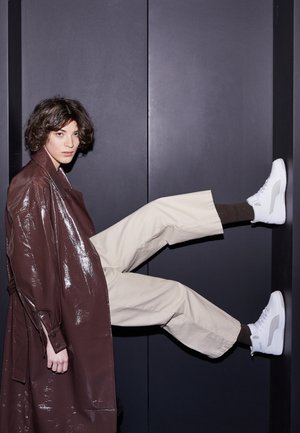 Trench-coat brun brillant porté sur un pantalon beige, mettant en valeur des baskets montantes blanches avec des découpes, sur un fond sombre.