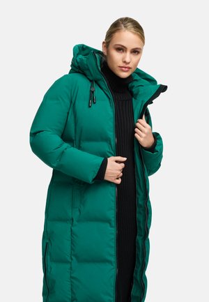 Long padded coat in vibrant green with a hood, featuring a front zipper, side pockets, and quilted texture. Black ribbed sweater underneath.