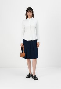 Vit långärmad skjorta med knappar som är kombinerad med en marinblå veckad kjol. Svarta loafers och en liten brun handväska tillför funktionella detaljer.