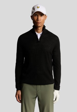 Pull noir à col zippé en tricot texturé, assorti à un pantalon vert olive et une casquette de baseball blanche arborant un logo jaune.