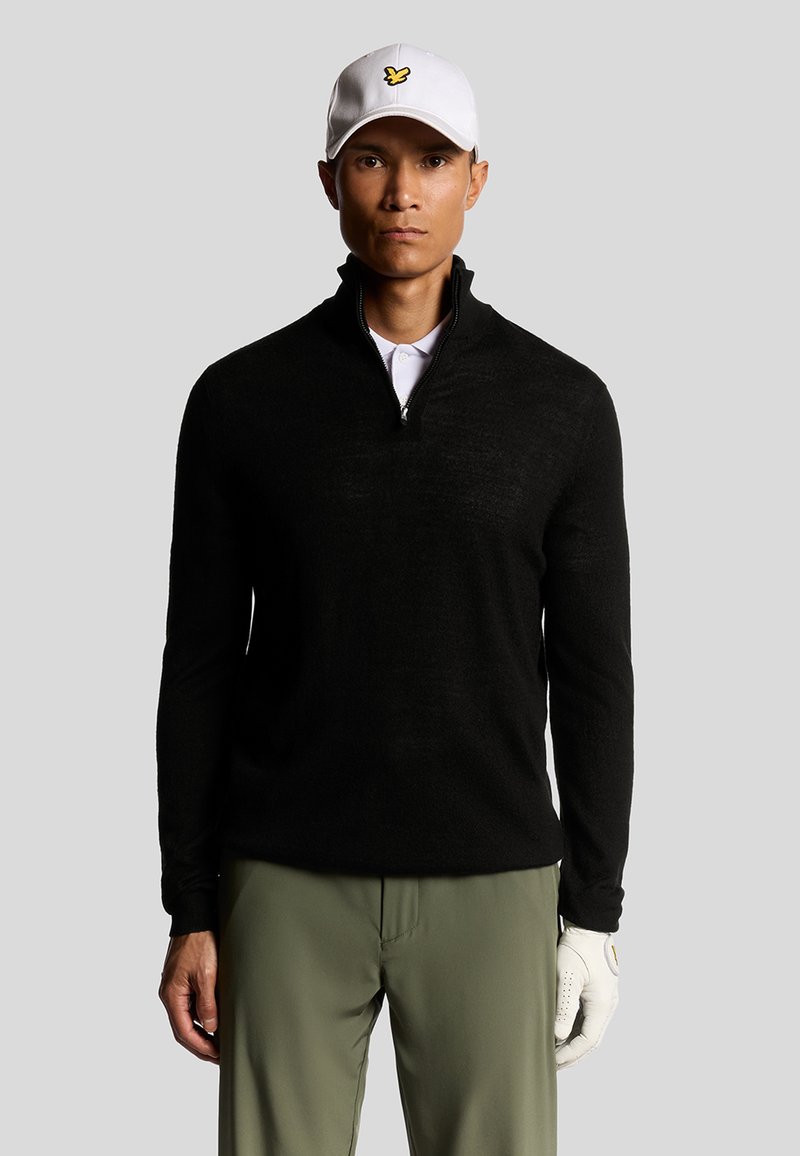 Maglione nero con zip e collo alto in maglia strutturata, abbinato a pantaloni verde oliva e un cappellino da baseball bianco con un logo giallo.