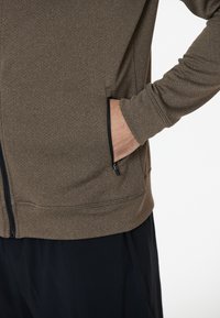 Veste brune zippée en tissu texturé, avec des poches latérales et une coupe ajustée. Les poignets sont serrés, créant une apparence élancée.