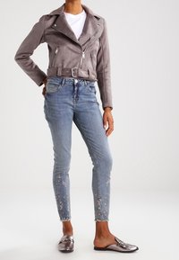 Veste en daim grise de motard avec fermeture éclair à l'avant et ceinture, associée à un jean bleu orné d'une broderie florale au niveau de l'ourlet et à des chaussures métalliques à enfiler.