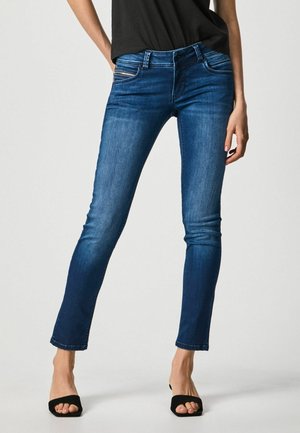Vrouw die nauwsluitende donkerblauwe jeans en zwarte open-toe hakken draagt, staand met licht gespreide benen tegen een effen achtergrond.