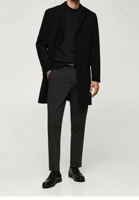 Manteau en laine noire porté sur une chemise noire, associé à un pantalon gris et des chaussures en cuir noires. Design simple et sur mesure avec une coupe ajustée.