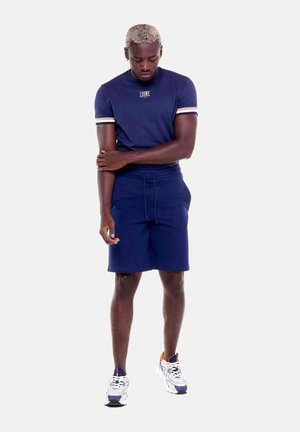 Man draagt marineblauw T-shirt en shorts met witte en oranje sneakers, staat en kijkt naar beneden terwijl hij zijn linkerarm vasthoudt.