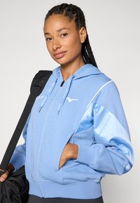 Sweatshirt azul claro com fecho éclair e textura suave, com mangas em satin de contraste, logótipo no peito e bolsos laterais. Saco de ginásio preto ao fundo.