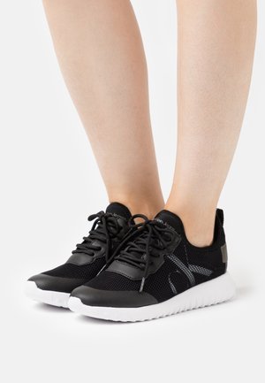 Baskets en mesh noir et cuir avec semelles blanches portées pieds nus, dotées d'une fermeture à lacets et d'un logo de marque discret sur les côtés.