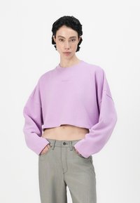 Lila lyhythihainen collegepaita, jossa on leveät hihat ja kirjailtu logo edessä, yhdistettynä korkeavyötäröisiin vaaleanharmaisiin housuihin, joissa on taskut.