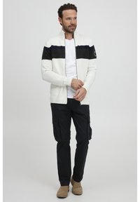 Cárdigan blanco de cremallera con franjas horizontales en azul marino y gris oscuro, llevado sobre una camiseta blanca, combinado con pantalones cargo negros y botas beige.