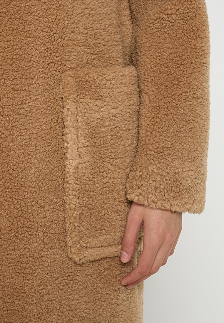 Manteau en polaire beige présentant une surface texturée et duveteuse, avec une poche à rabat et une coupe ample. Les extrémités des manches sont également texturées.