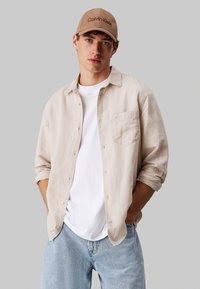 Camisa de lino beige de manga larga, camiseta blanca debajo, jeans azul claro y gorra marrón de Calvin Klein con logotipo bordado.