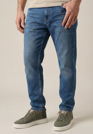 Mann trägt blaue Slim-Fit-Jeans und graue Schnürschuhe, steht vor neutralem Hintergrund.