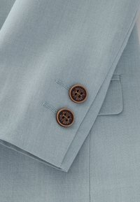 Lichtblauw, op maat gemaakt stof met een gestructureerde weving, met twee bruine knopen gegraveerd met "AUTHENTIC" naast een net zakje.