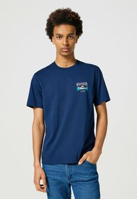Marineblå bomulds-t-shirt med korte ærmer, prydet med et grafisk logo på venstre bryst. Sammen med mørke denim jeans.