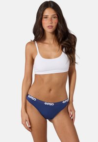 Femme aux longs cheveux foncés portant un brassière de sport blanche et un sous-vêtement bleu marine avec une ceinture blanche à logo PSD, debout devant un fond uni.