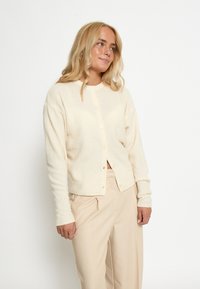 Beige adīta jaka ar apaļu kakla izgriezumu, priekšpuses pogām un ribotām piedurknēm. Saskaņota ar gaiši bežām biksēm. Mīksta tekstūra, ikdienišķs dizains.