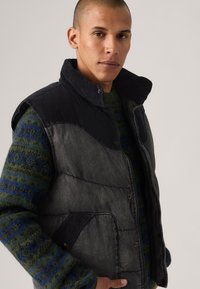 Chaleco acolchado en gris oscuro y negro de pana, con un cuello alto, botones de presión y bolsillos laterales. Lleva puesto sobre un suéter verde con estampado.