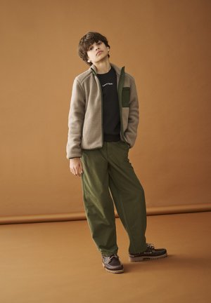 Una giovane persona è in piedi davanti a uno sfondo marrone indossando una giacca in pile beige, una camicia nera, pantaloni verde oliva e stivali marroni.