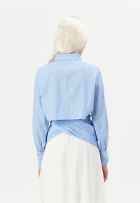 Blå og hvit stripet bluse med pynt, lange ermer og knyttet bakdesign, sammen med en hvit teksturert skjørt.