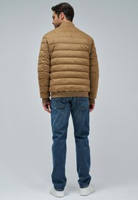 Chaqueta acolchada marrón con costuras horizontales y puños acanalados, combinada con jeans azules. La chaqueta tiene un cuello alto y es ligera.
