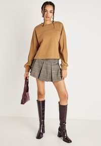 Un pulover scurt maro cu detalii de cusătură, o fustă plisată gri cu carouri, o geantă burgundy și cizme lucioase burgundy până la genunchi.