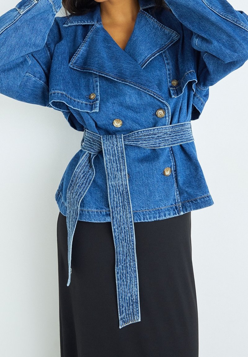 Jeansjacka i mellanblå nyans, med dubbelknapprad i fronten, stora slag och midjebälte i samma tyg. Strukturerad med synliga sömmar.