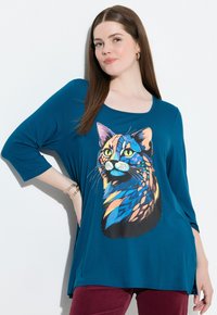 Camiseta holgada en color azul verdoso que presenta un gráfico colorido de la cara de un gato con patrones intrincados. Tela suave con escote redondo y mangas de 3/4.