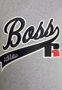 Tissu gris avec un texte brodé en noir et blanc "Boss Athletic" et un patch logo stylisé en noir et rouge sur le côté inférieur droit.