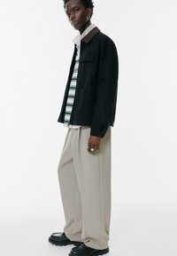 Schwarze Jacke mit braunem Kragen, über einem gestreiften Hemd getragen. Breite beigefarbene Hosen und schwarze Schuhe vervollständigen das Outfit. Silberne Ringe als Akzente.