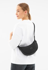 Borsa a tracolla nera in nylon con forma curva, cinturino regolabile e patch con logo; indossata sopra una camicia bianca e visibile di lato.