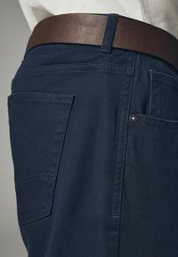 Dunkelblaue Stoffhosen verfügen über eine dezente gerippte Textur, einem braunen Ledergürtel und mehreren Taschen mit sichtbaren Nähdetails.