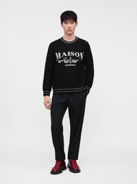 Νέος άντρας φοράει μαύρο πουλόβερ Maison Margiela, μαύρο παντελόνι και κόκκινα δερμάτινα μποτάκια με κορδόνια, στέκεται μπροστά σε απλό λευκό φόντο.