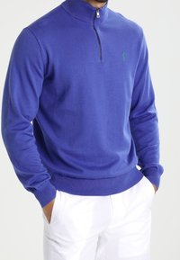 Pull bleu royal avec un col haut et une fermeture éclair, doté de poignets et d'un ourlet côtelés, mettant en valeur un logo vert sur la poitrine.